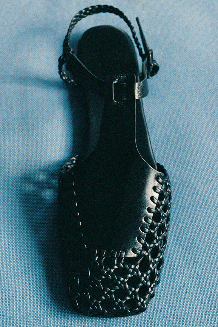 A black square-toed sandals with a t-bar strap and hand-woven design, featuring a combination of veg-tanned calf leather and supple lamb skin, with a rubber heel.
Photographed on a blue background.