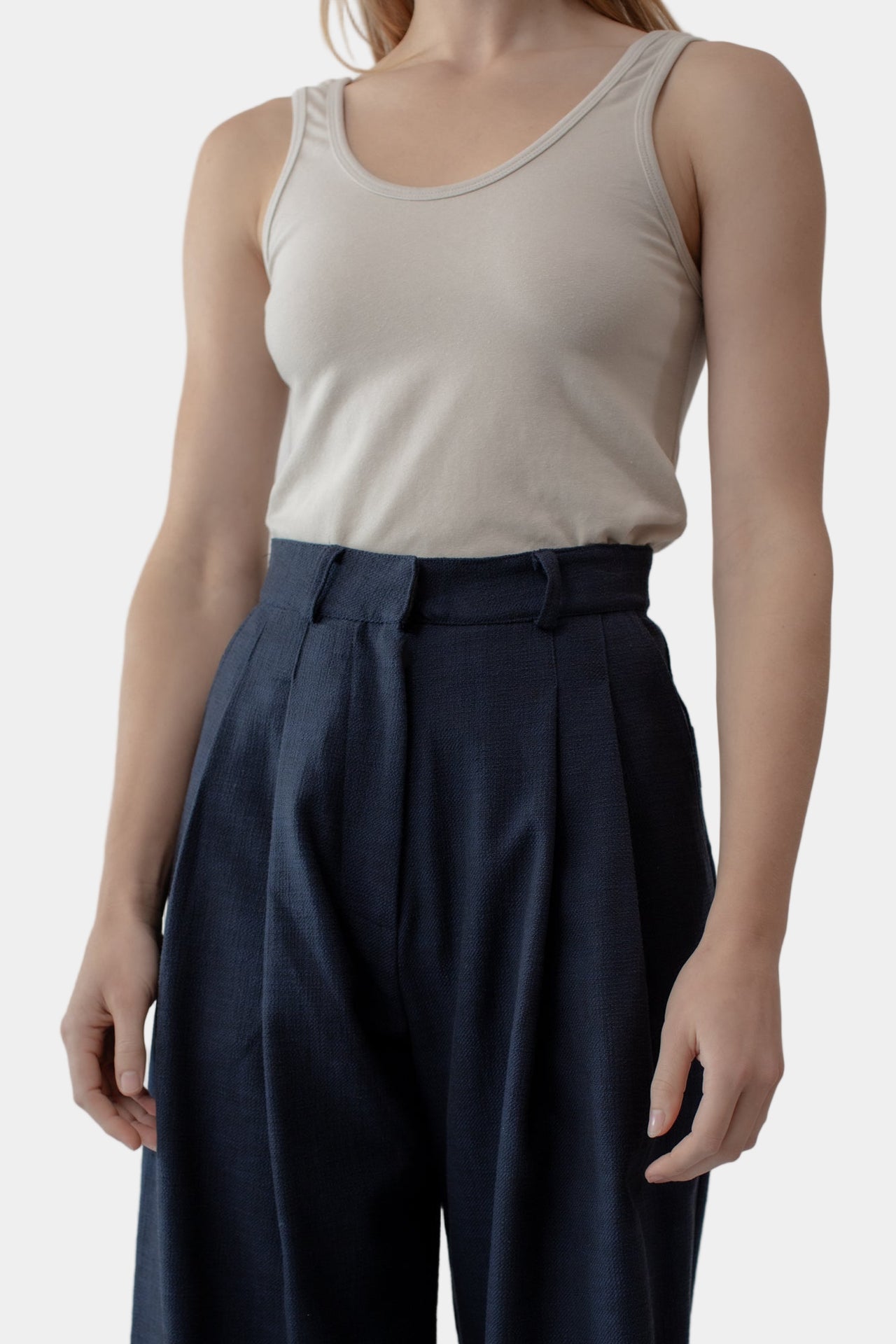 A woman stands against a white background wearing navy high-waisted trousers with a double front pleat and side entry pockets. She has on a beige tank top and is posing by adjusting the waistband of the trousers.
