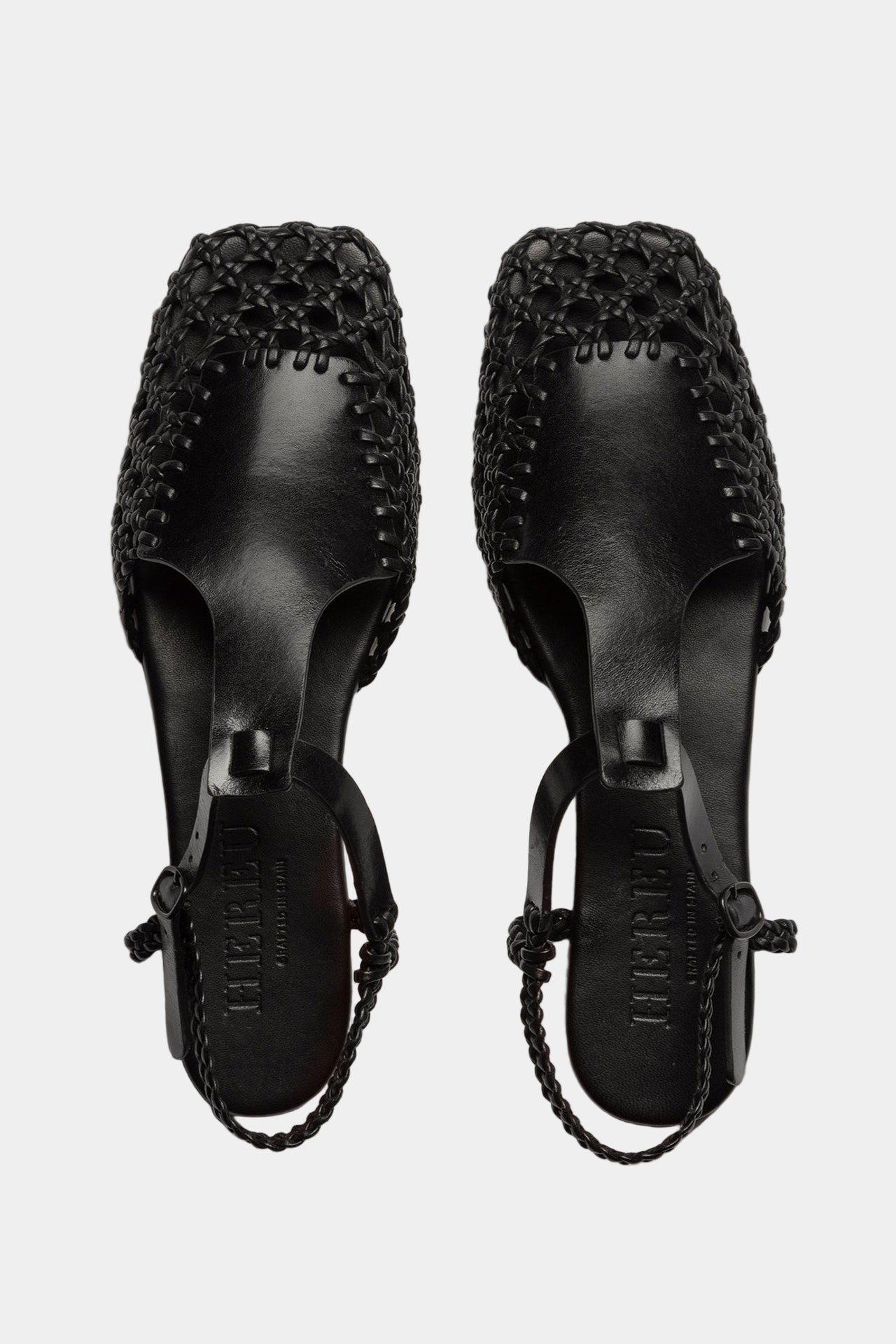 A pair of black square-toed sandals with a t-bar strap and hand-woven design, featuring a combination of veg-tanned calf leather and supple lamb skin, with a rubber heel.