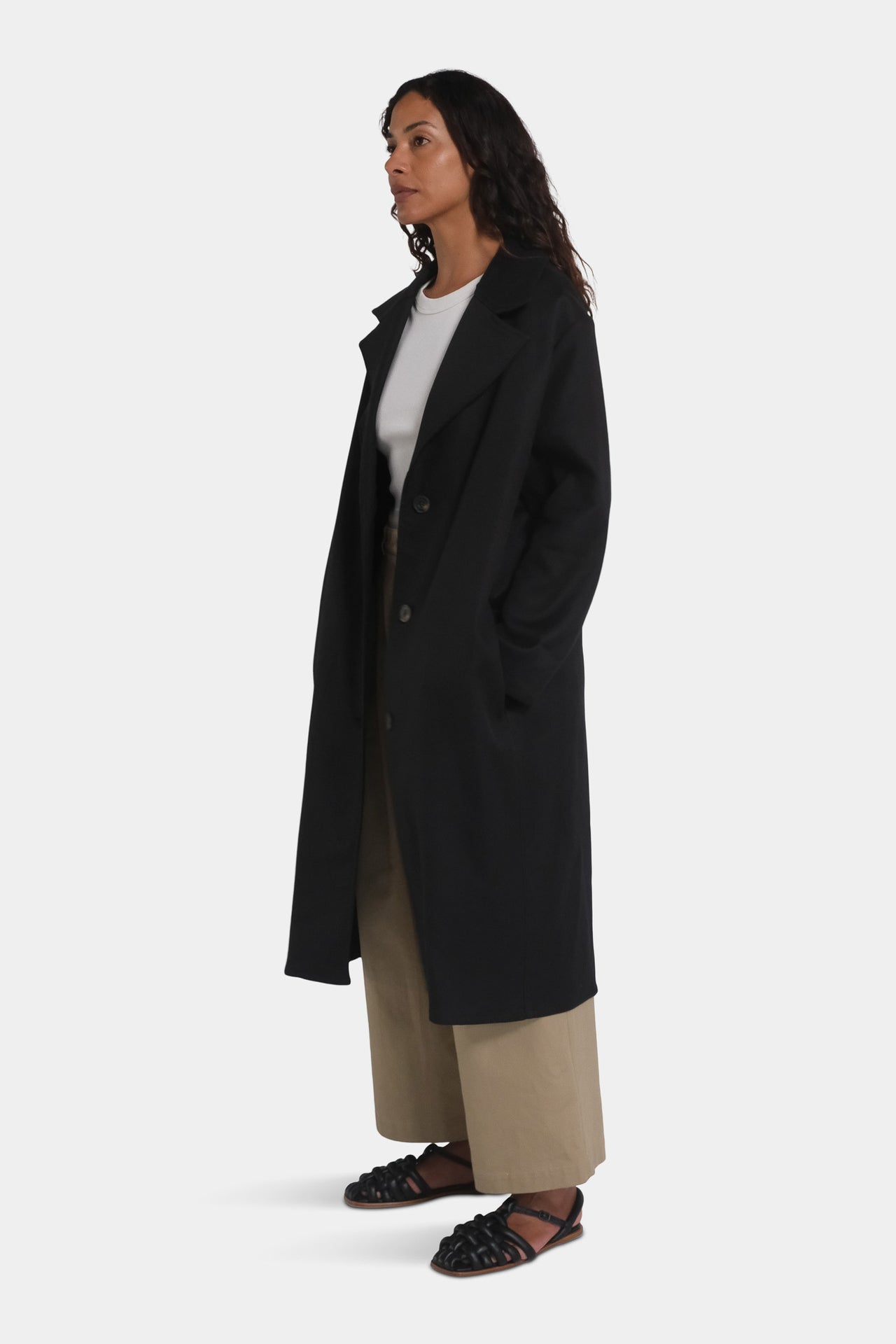 A female model wearing a long black overcoat with notched lapels and a mid-calf length, paired with a white top and beige trousers.