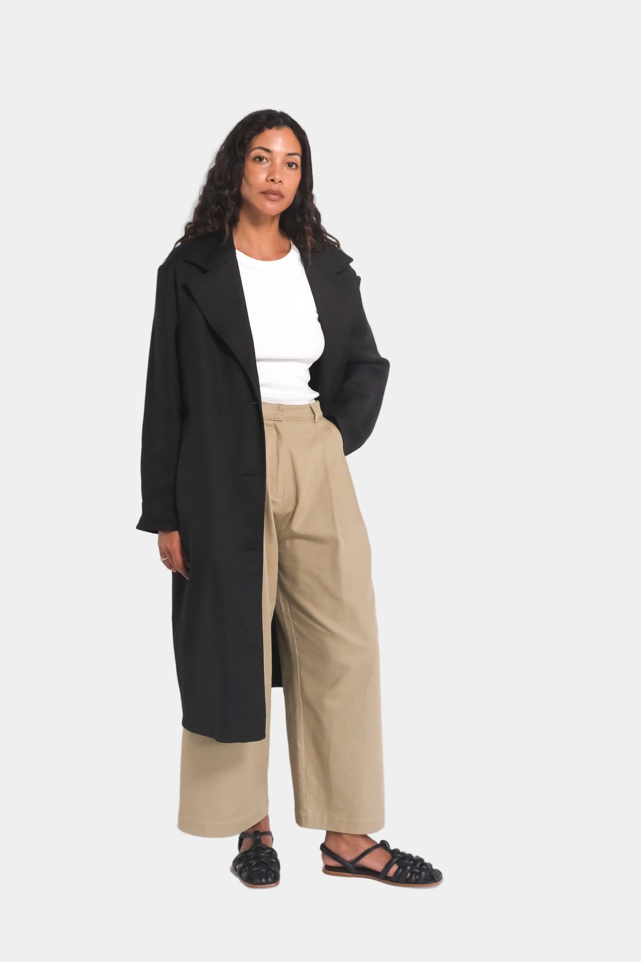 A female model wearing a long black overcoat with notched lapels and a mid-calf length, paired with a white top and beige trousers.
