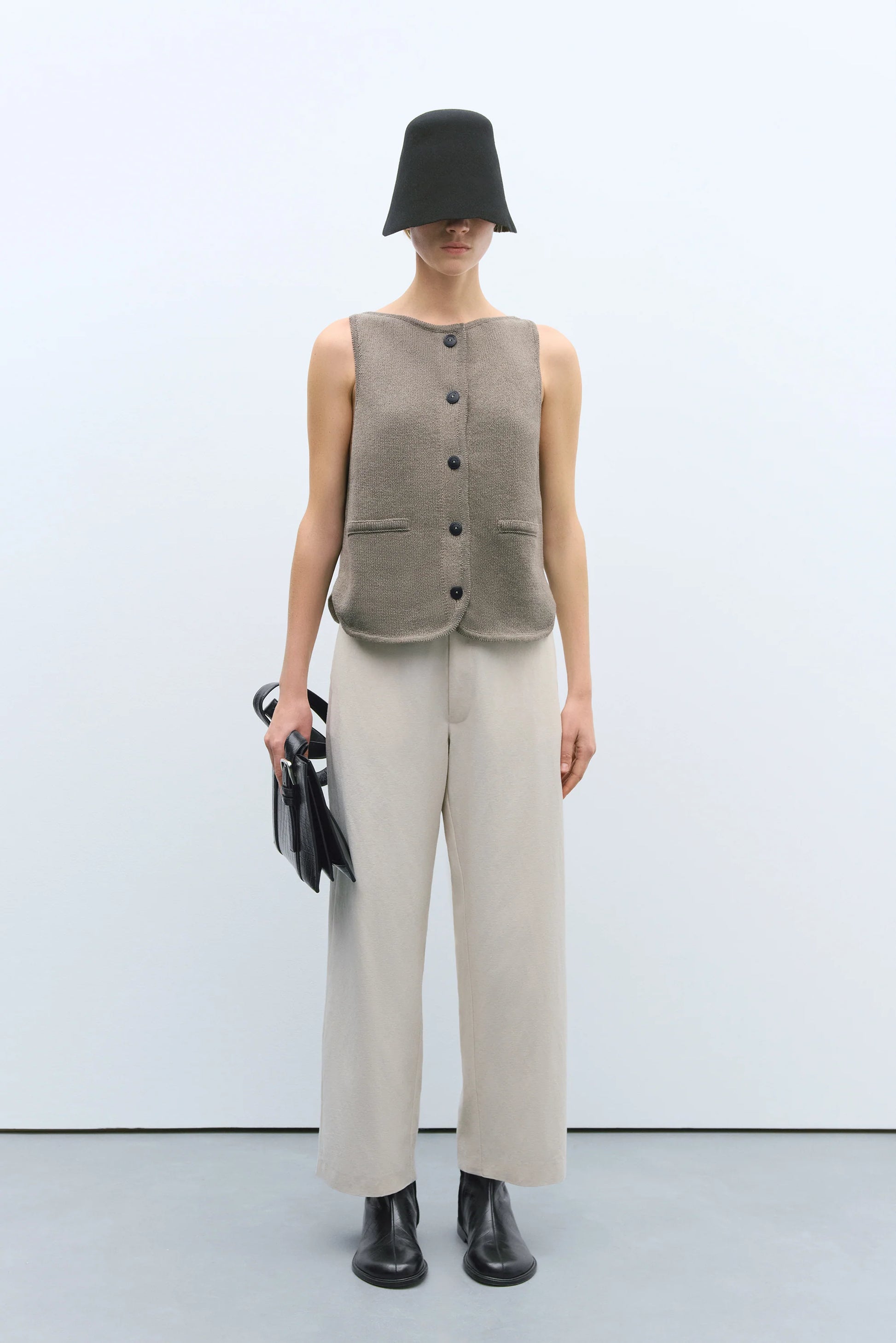 Person wearing a beige sleeveless vest, white pants, and black shoes on a white background