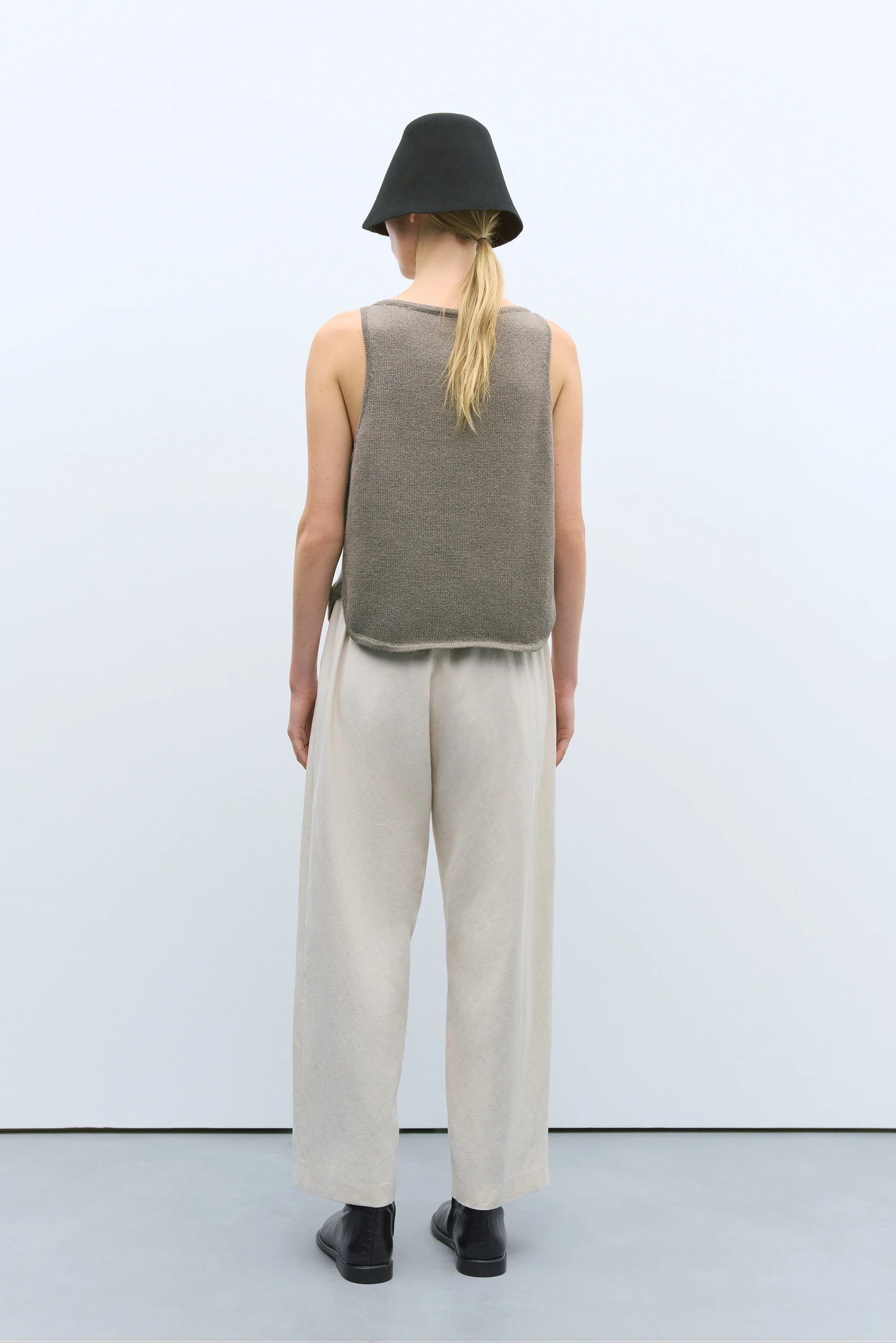 Person wearing a sleeveless green top and beige pants with a black hat on a white background