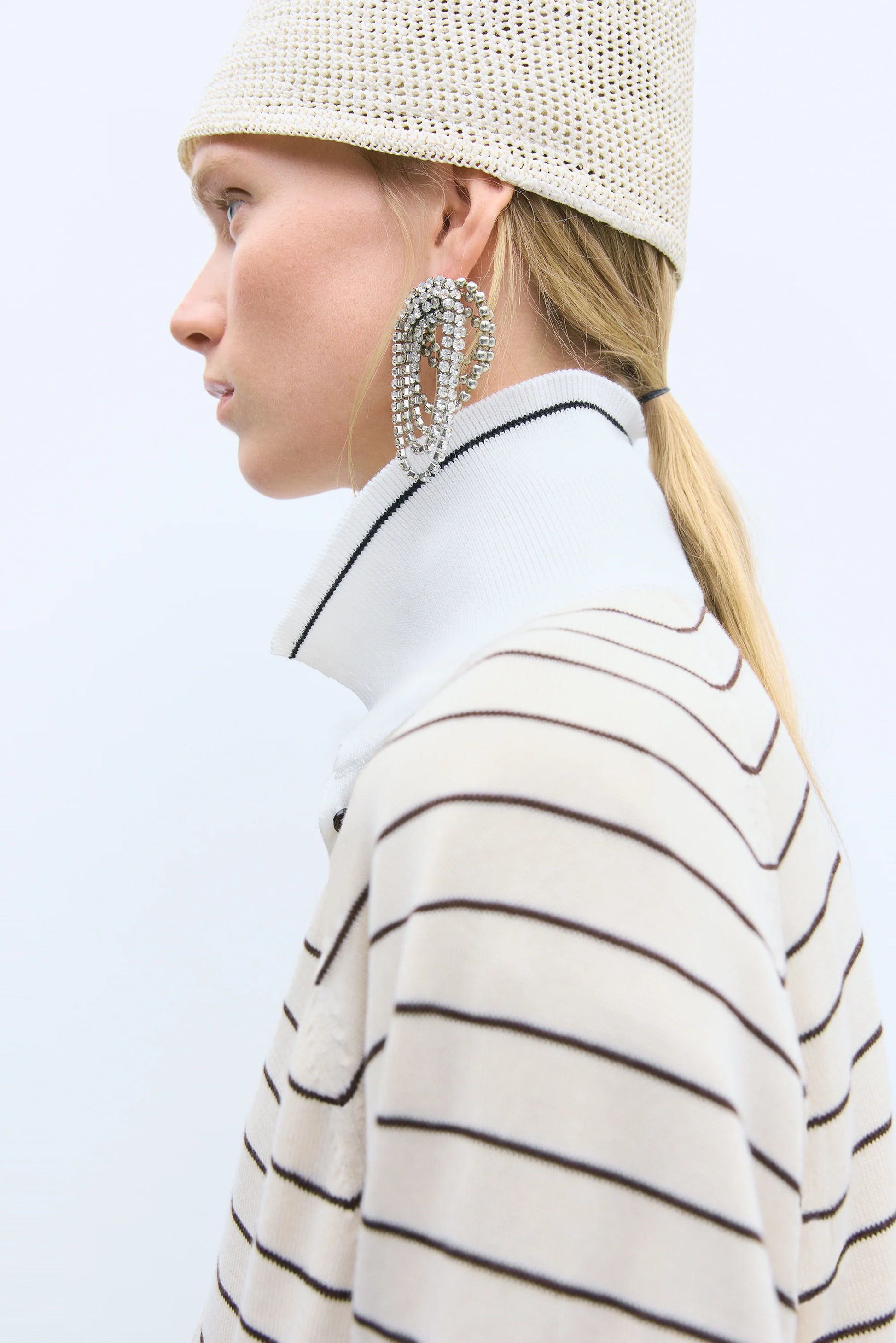 Person wearing a striped coat, white beanie, and large hoop earrings on a light background