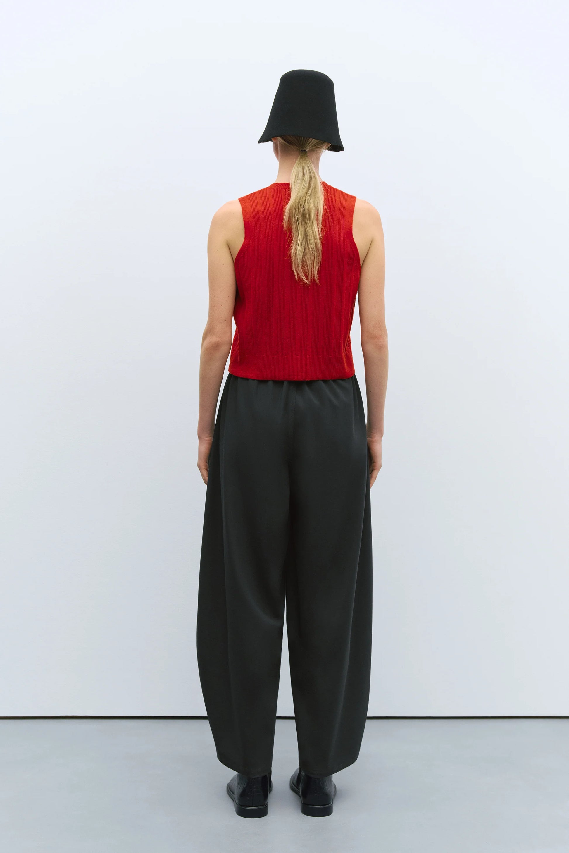 Person wearing a red sleeveless top and gray wide-leg pants with a black hat on a white background