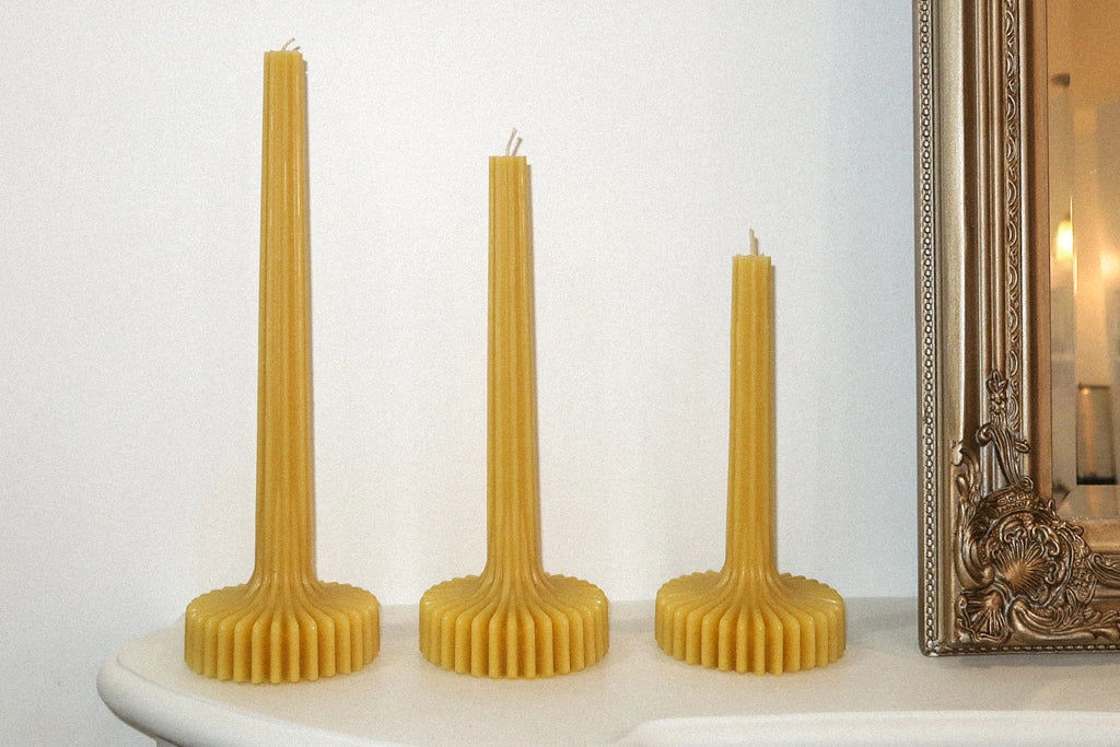 Three tall, ribbed beeswax candles on a white surface with a decorative mirror in the background.