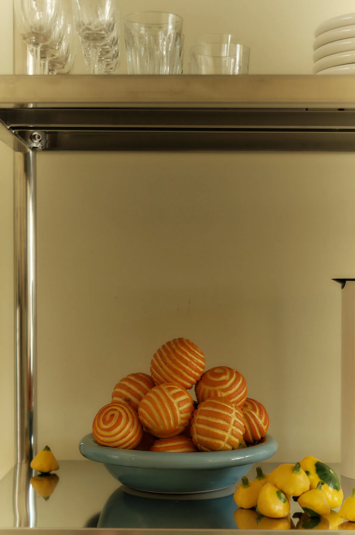 oranges in a bowl sitting on stainless steal shelves with glasses and plates.