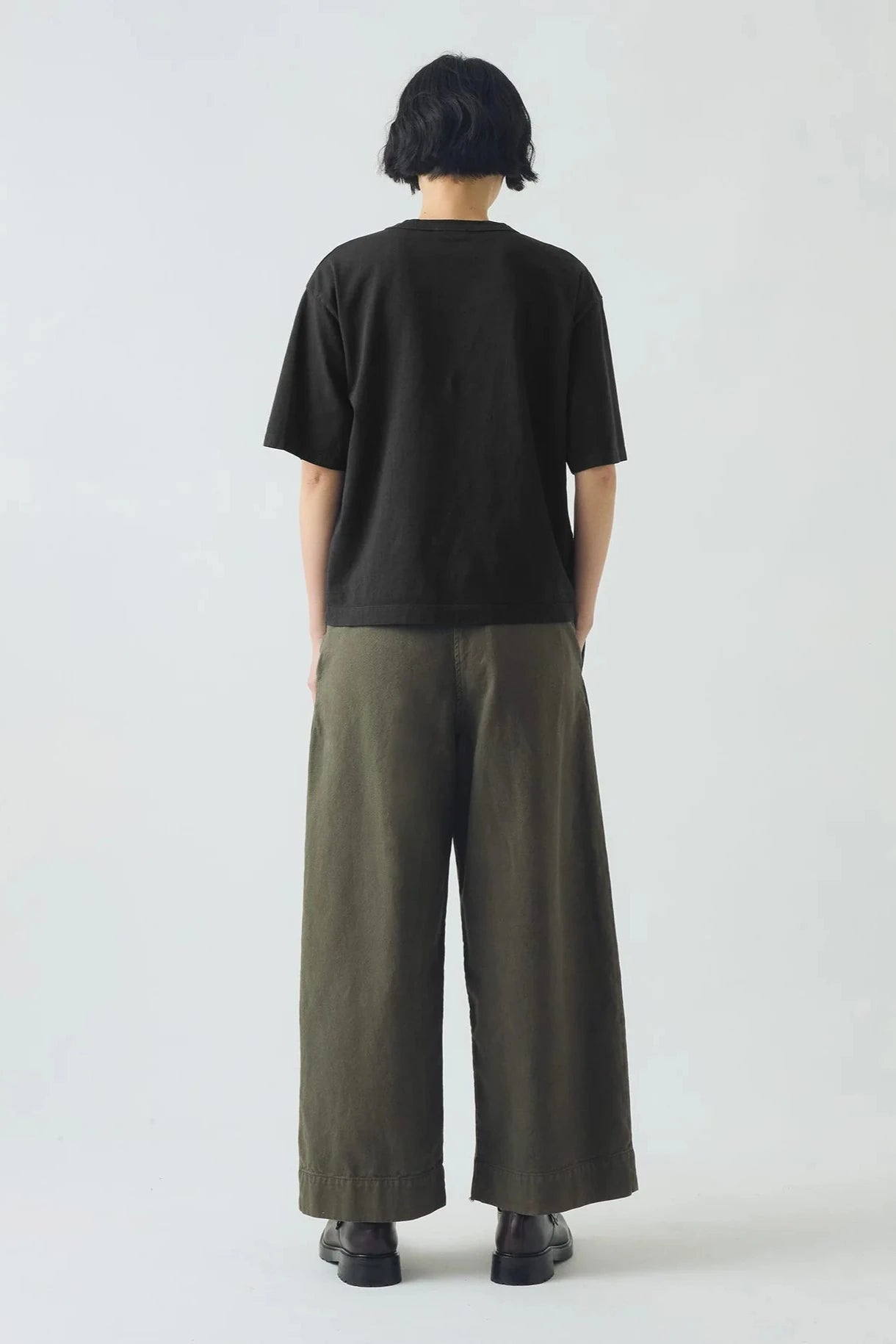 Person wearing a black t-shirt and olive green pants on a white background, viewed from the back.