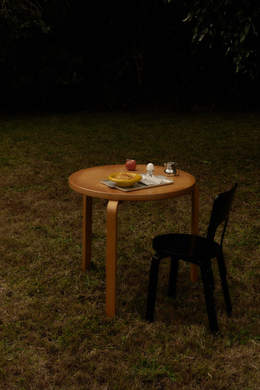 Round wooden table and a chair on a grassy field, with a silver tray and mustard breakfast bowl.