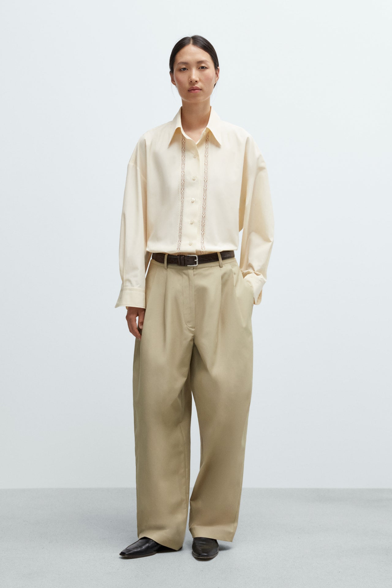 A women wearing an oversized cream shirt with lace details on a plain background