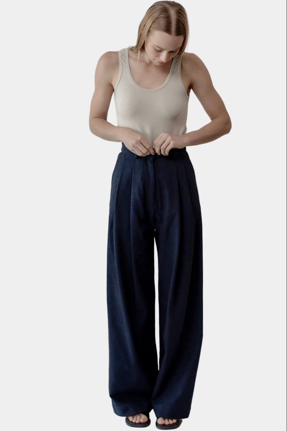 A woman stands against a white background wearing navy high-waisted trousers with a double front pleat and side entry pockets. She has on a beige tank top and is posing by adjusting the waistband of the trousers.