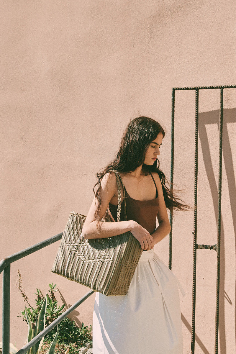 Woman holding a woven bag against a pink wall with a metal gate.
