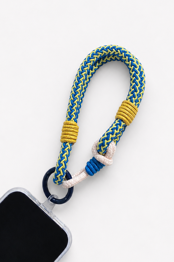 A braided rope phone strap in blue, yellow, and white, attached to a black ring and shown with part of a smartphone on a clean white background - East Coast General