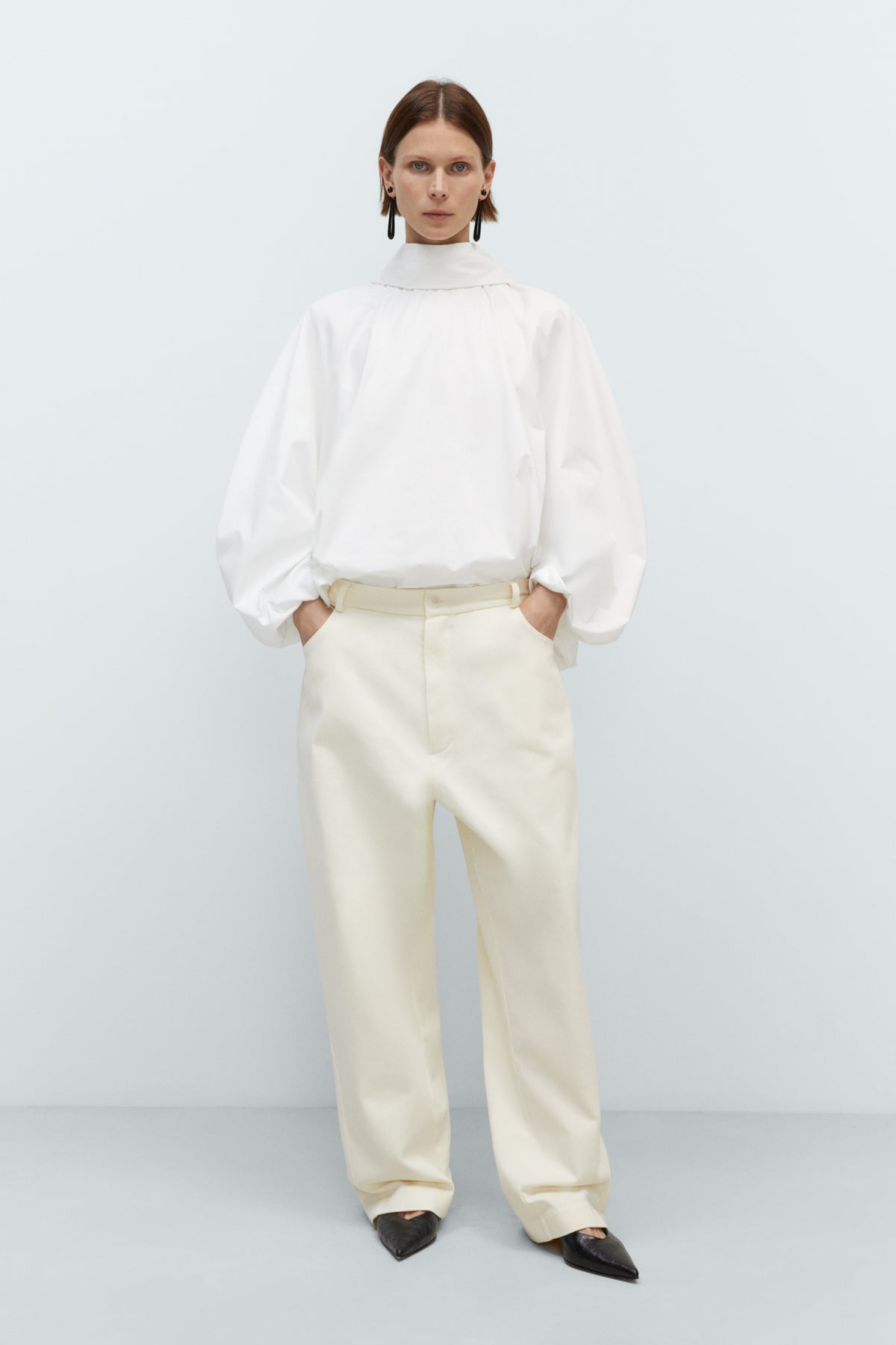 Person wearing a white outfit with a high collar and beige pants on a light gray background