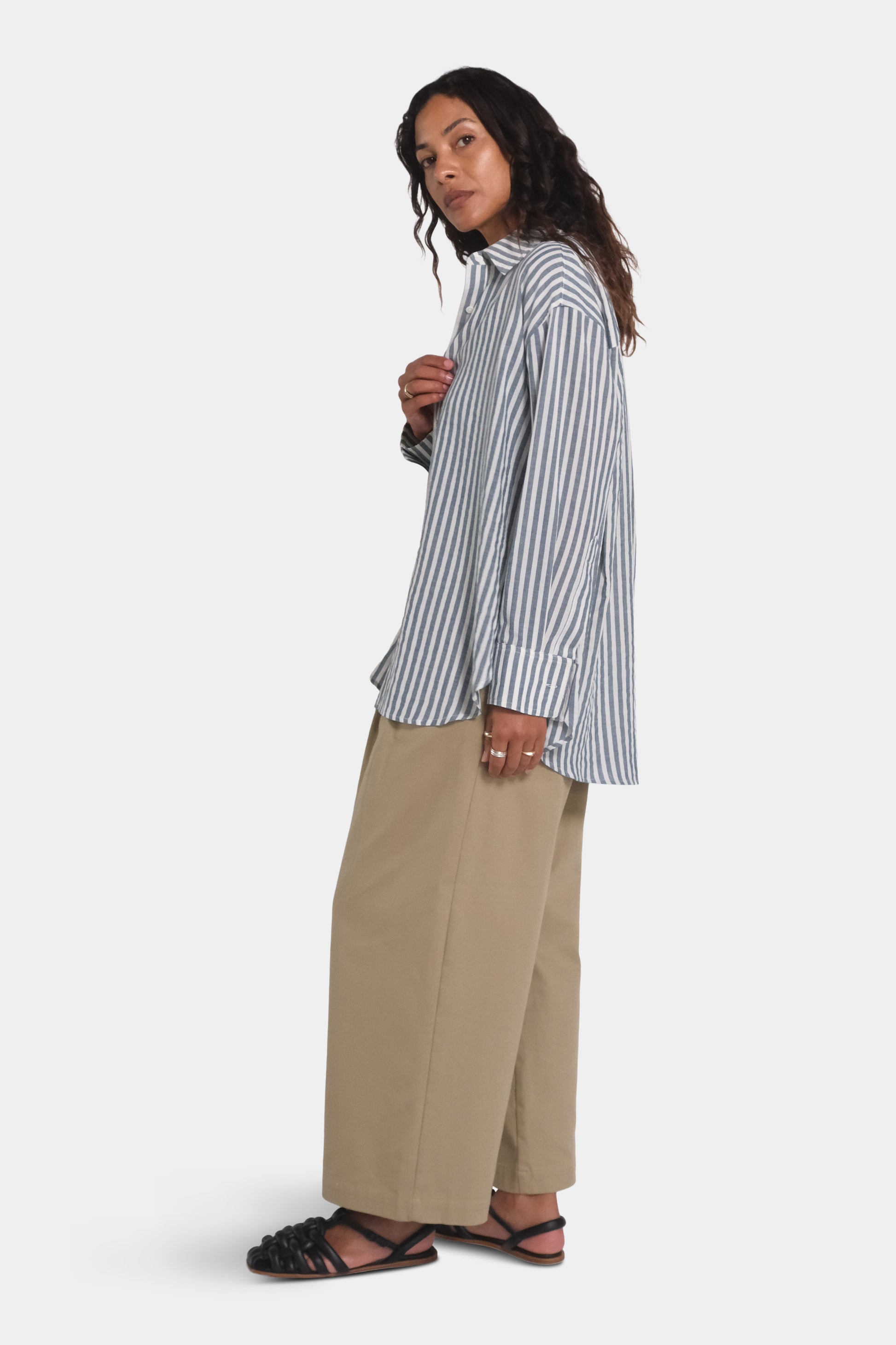 a women wearing a stripe blue and white shirt and beige trousers, viewed from the side on a plain background