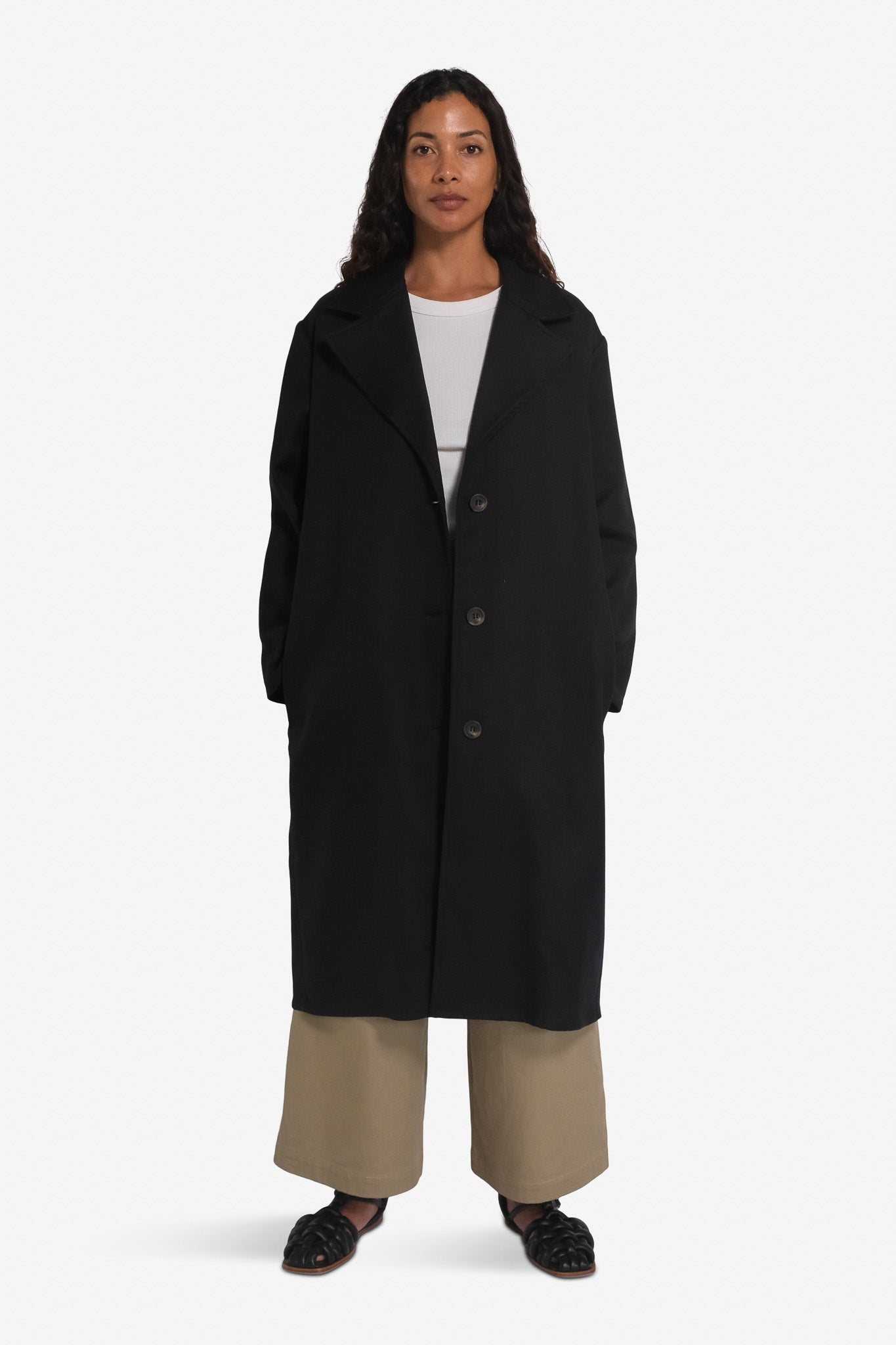 A female model wearing a long black overcoat with notched lapels and a mid-calf length, paired with a white top and beige trousers.