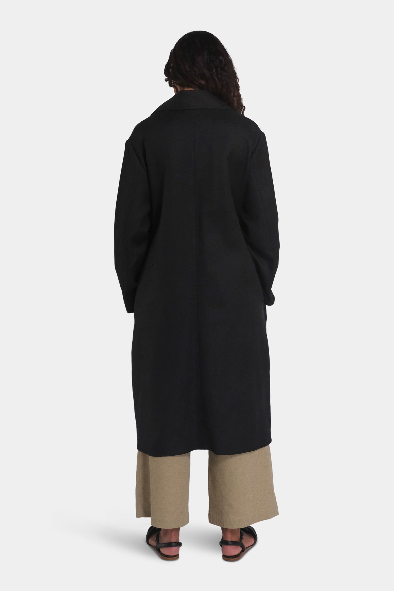 A female model wearing a long black overcoat falling to a mid-calf length, paired with beige trousers. Viewed from the back