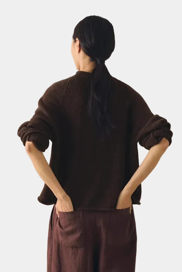 Person wearing a brown sweater and pants standing against a beige curtain.