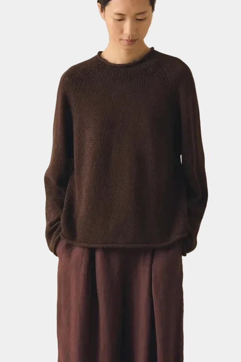 Person wearing a brown outfit standing against a beige curtain.