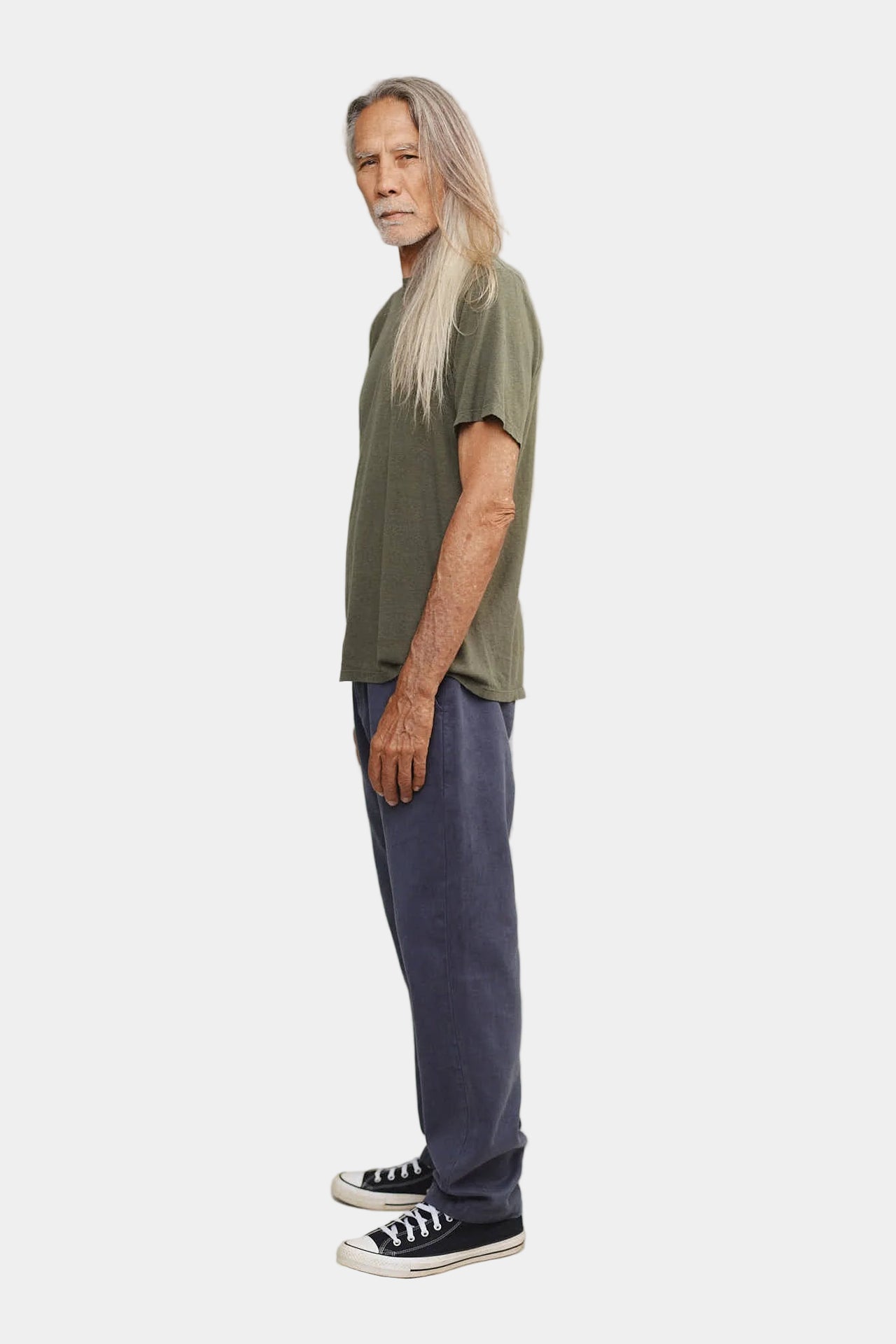 Person wearing an olive green shirt and blue pants with black shoes on a plain background