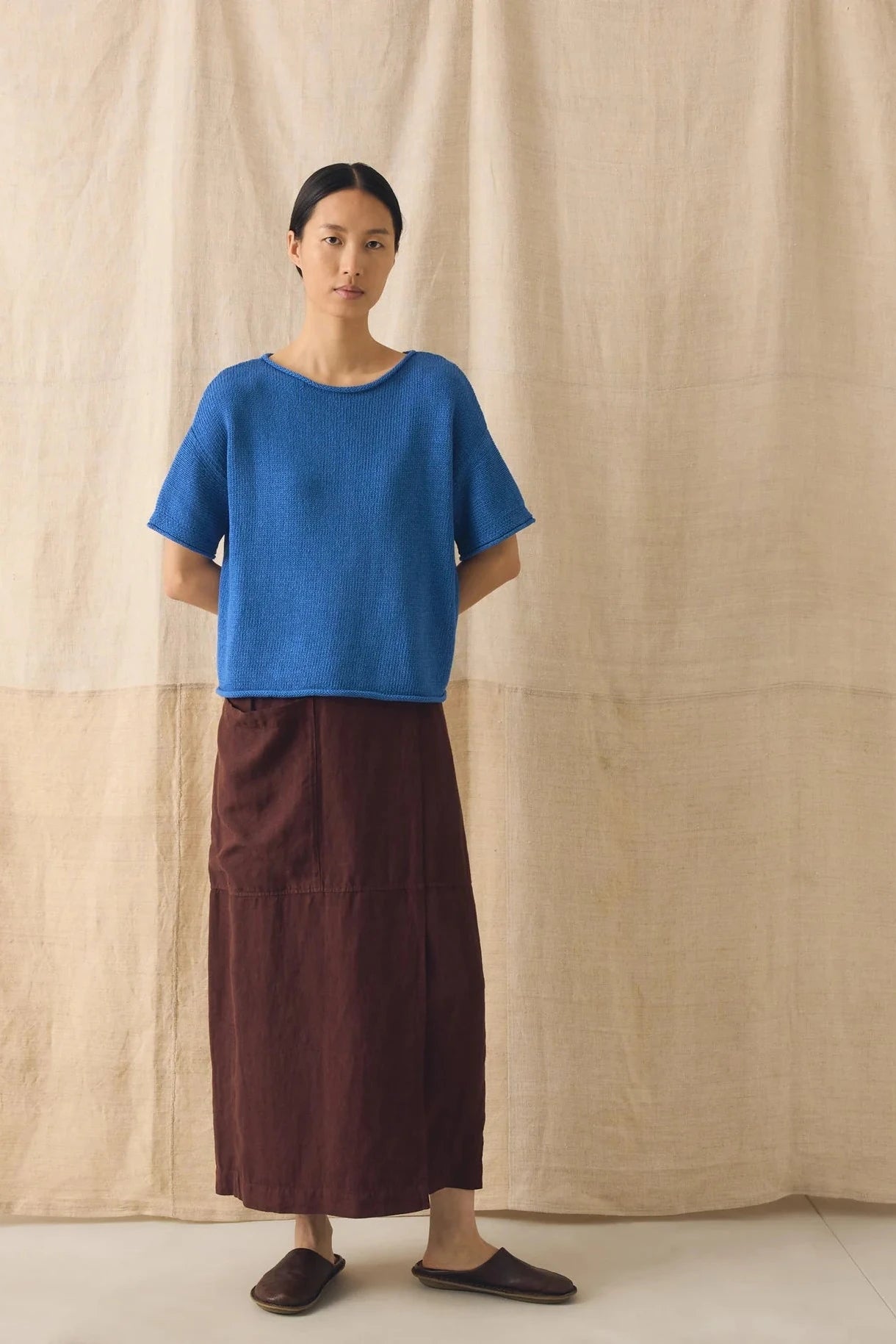 Person wearing a blue top and brown skirt standing against a beige curtain.