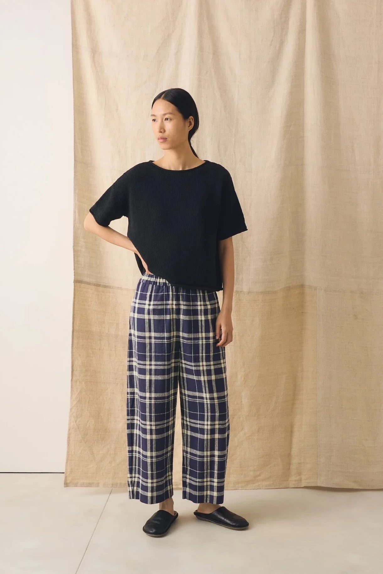 Person wearing a black top and blue plaid pants standing against a beige curtain.