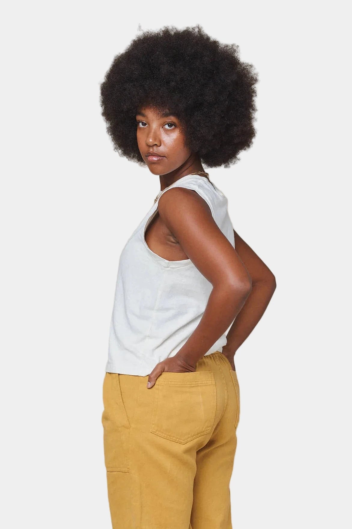 Woman wearing a white tank top and mustard yellow pants against a beige background
