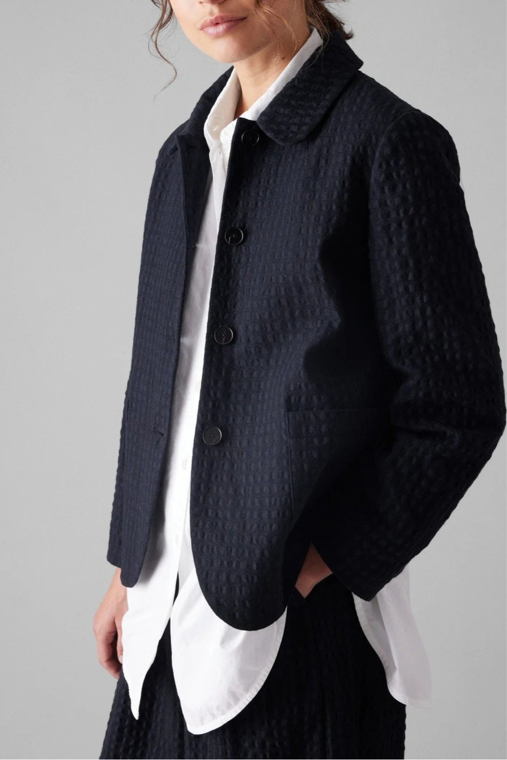 Woman wearing a dark textured jacket over a white shirt on a plain background