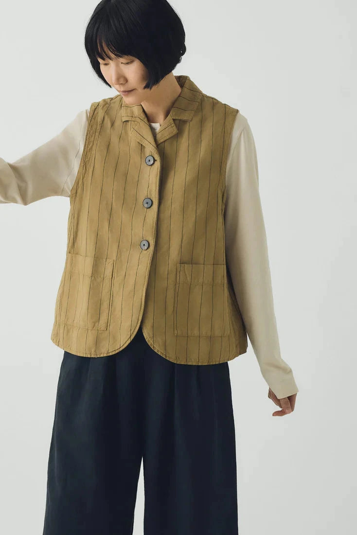 Person wearing a beige vest over a cream shirt with dark pants on a white background