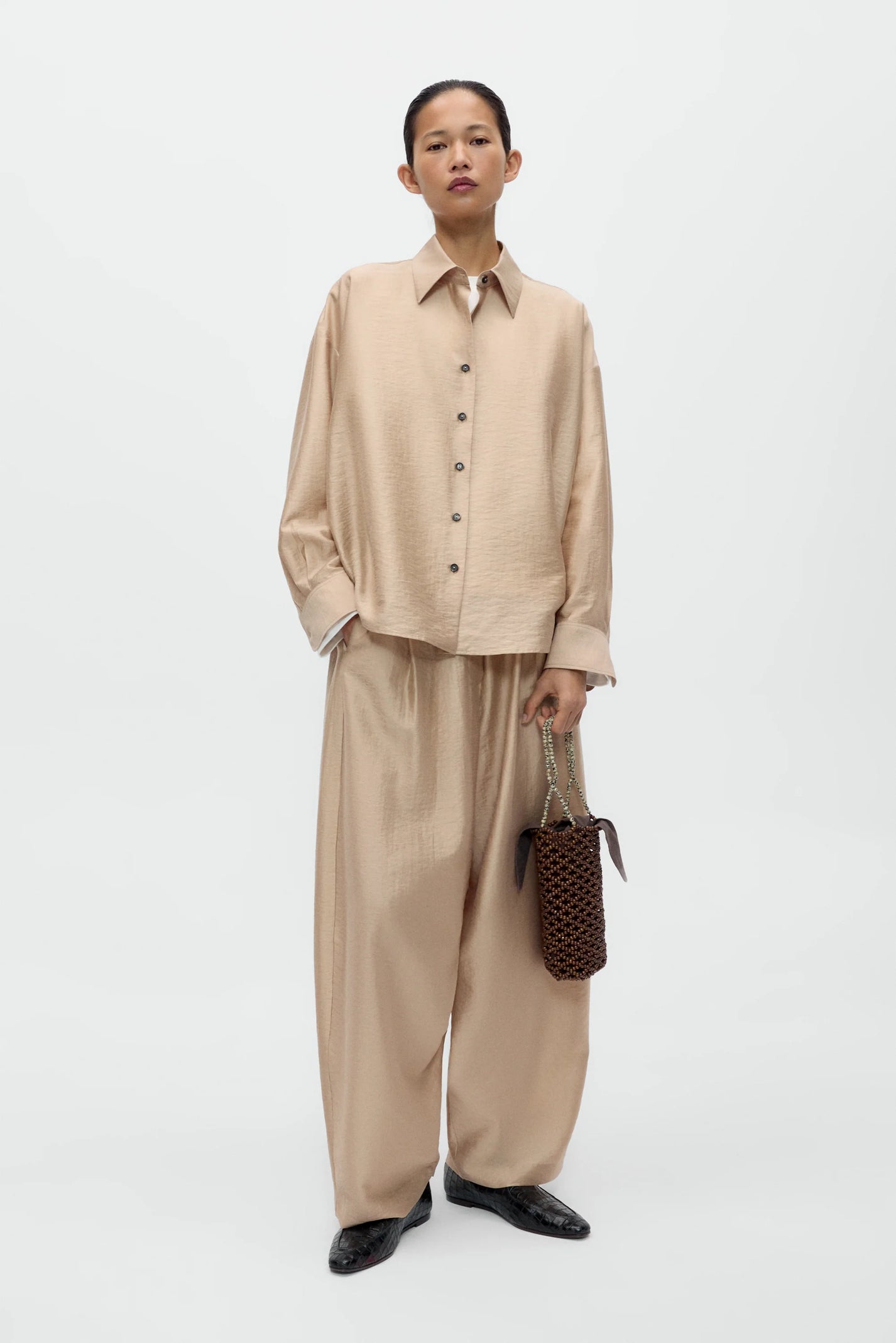 Person wearing a beige shirt and pants holding a woven bag on a white background