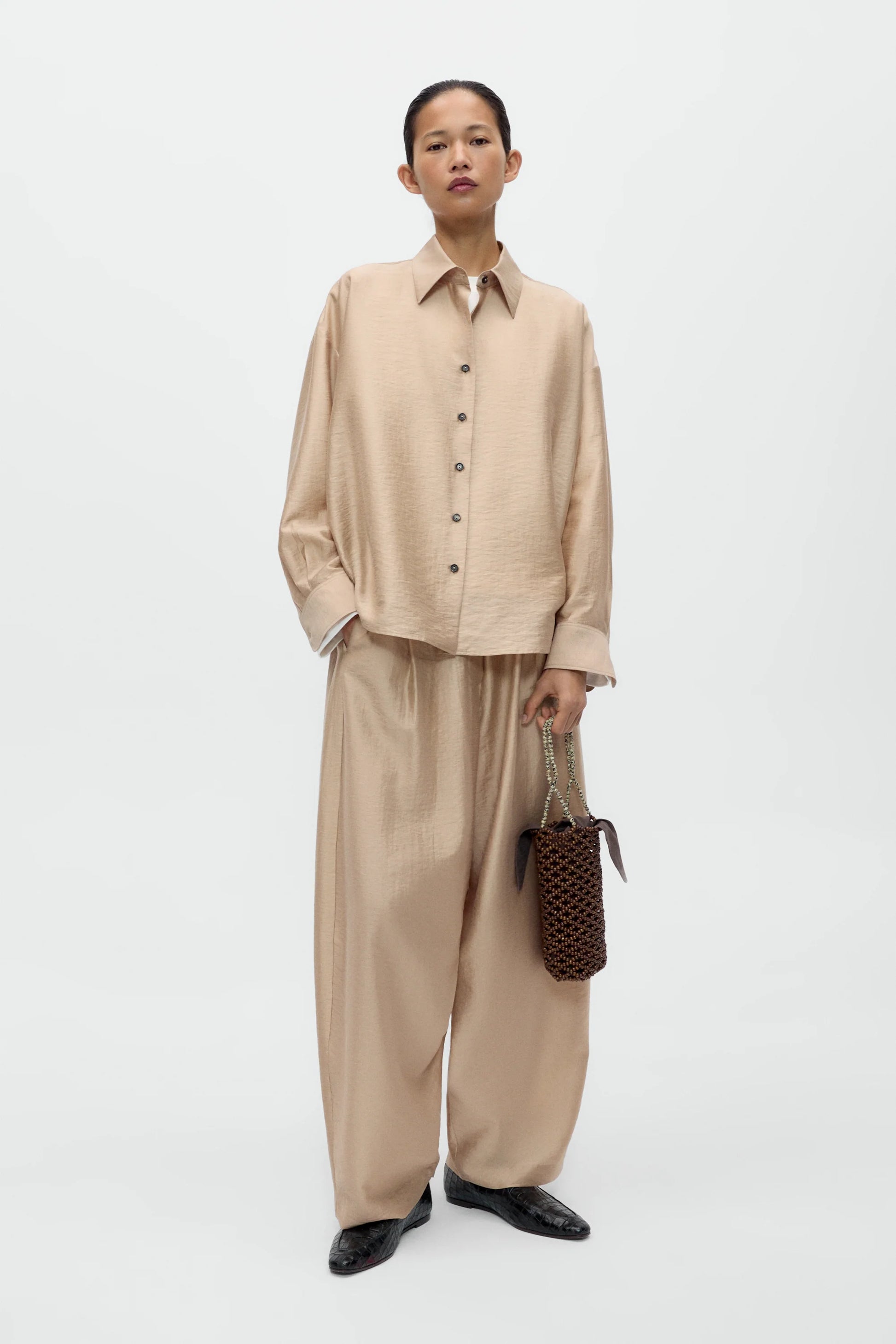 Person wearing a beige shirt and pants holding a woven bag on a white background