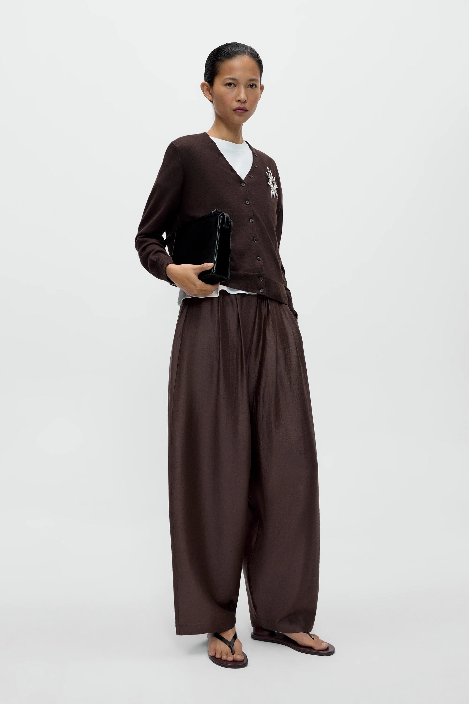 A women wearing a brown cardigan and white t-shirt with brown maxi pants, standing against a white background