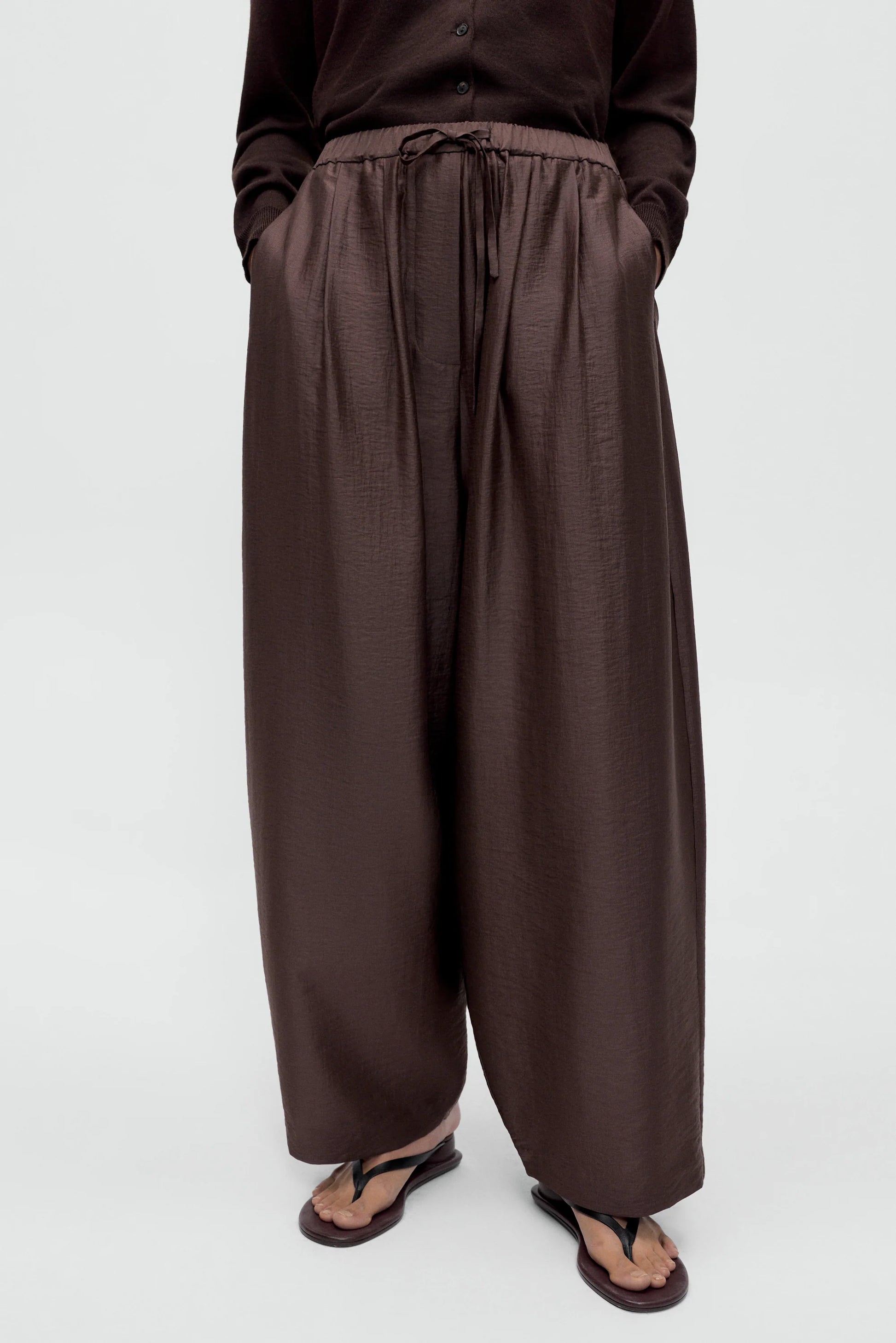 A women wearing a brown cardigan with brown maxi pants, standing against a white background, styled with brown sandals