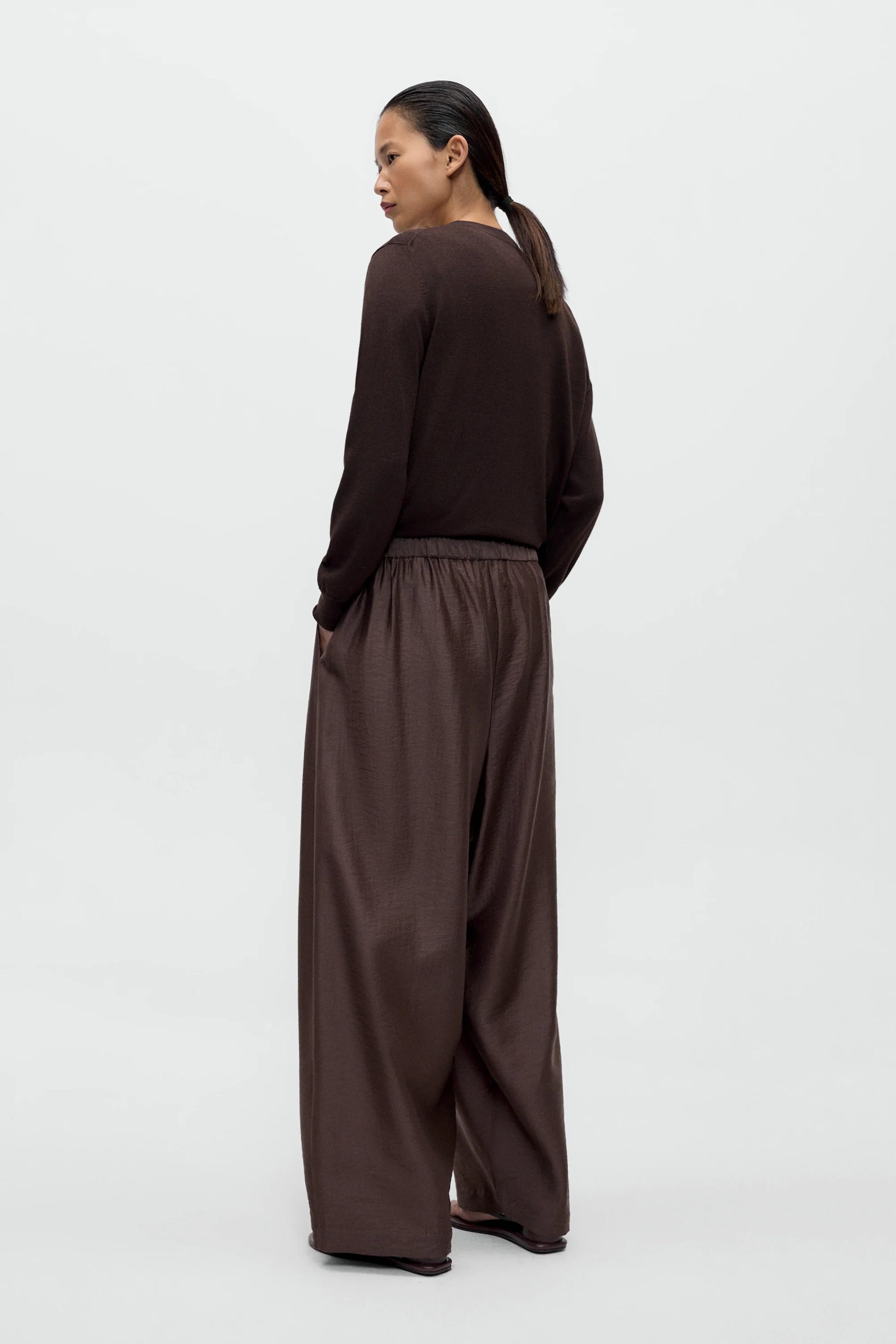 A women wearing a brown cardigan and white t-shirt with brown maxi pants, standing against a white background, back view