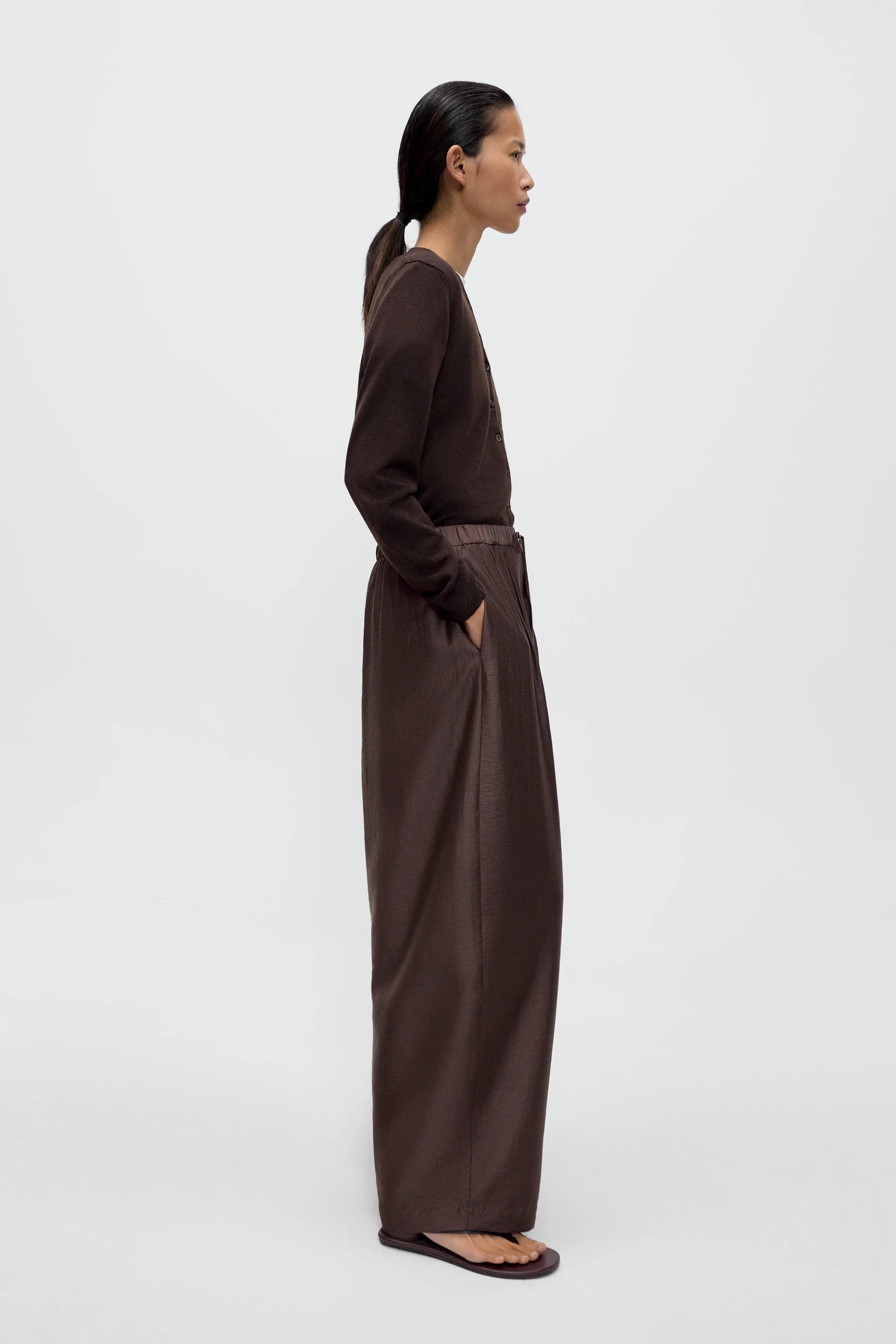 A women wearing a brown cardigan and white t-shirt with brown maxi pants, standing against a white background, view from the side