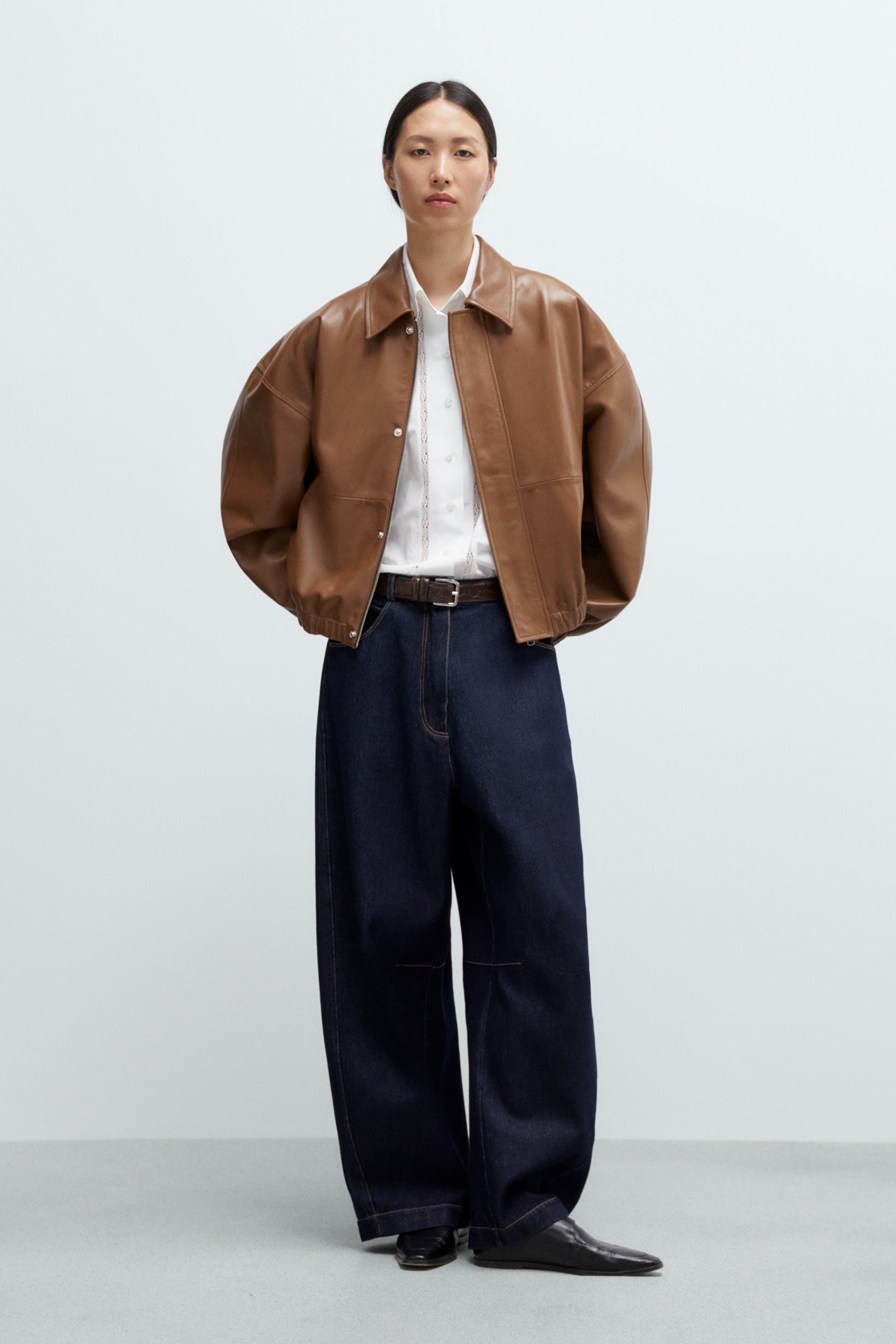 Person wearing a brown jacket, white shirt, and dark blue curved leg jeans on a light gray background