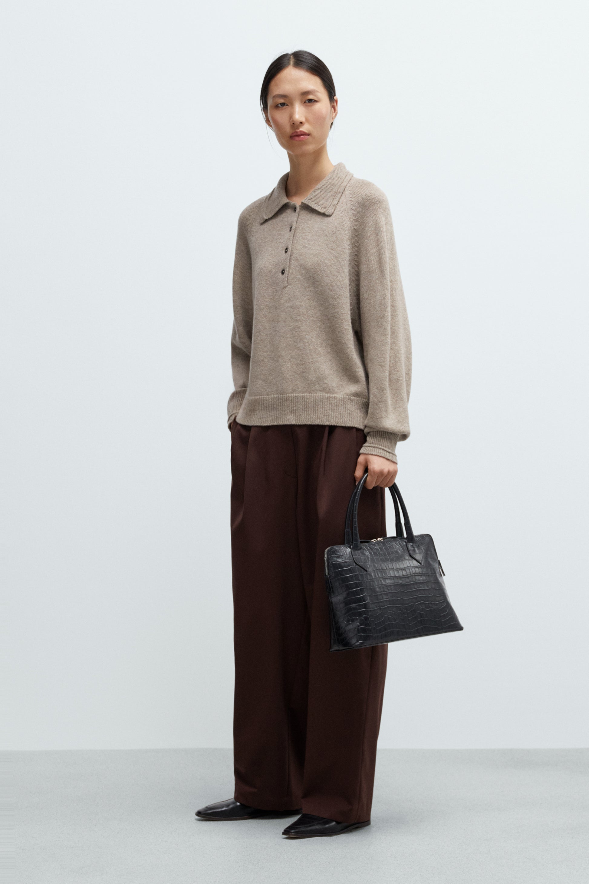 Woman wearing a beige polo collared sweater and brown pants holding a black handbag on a white background
