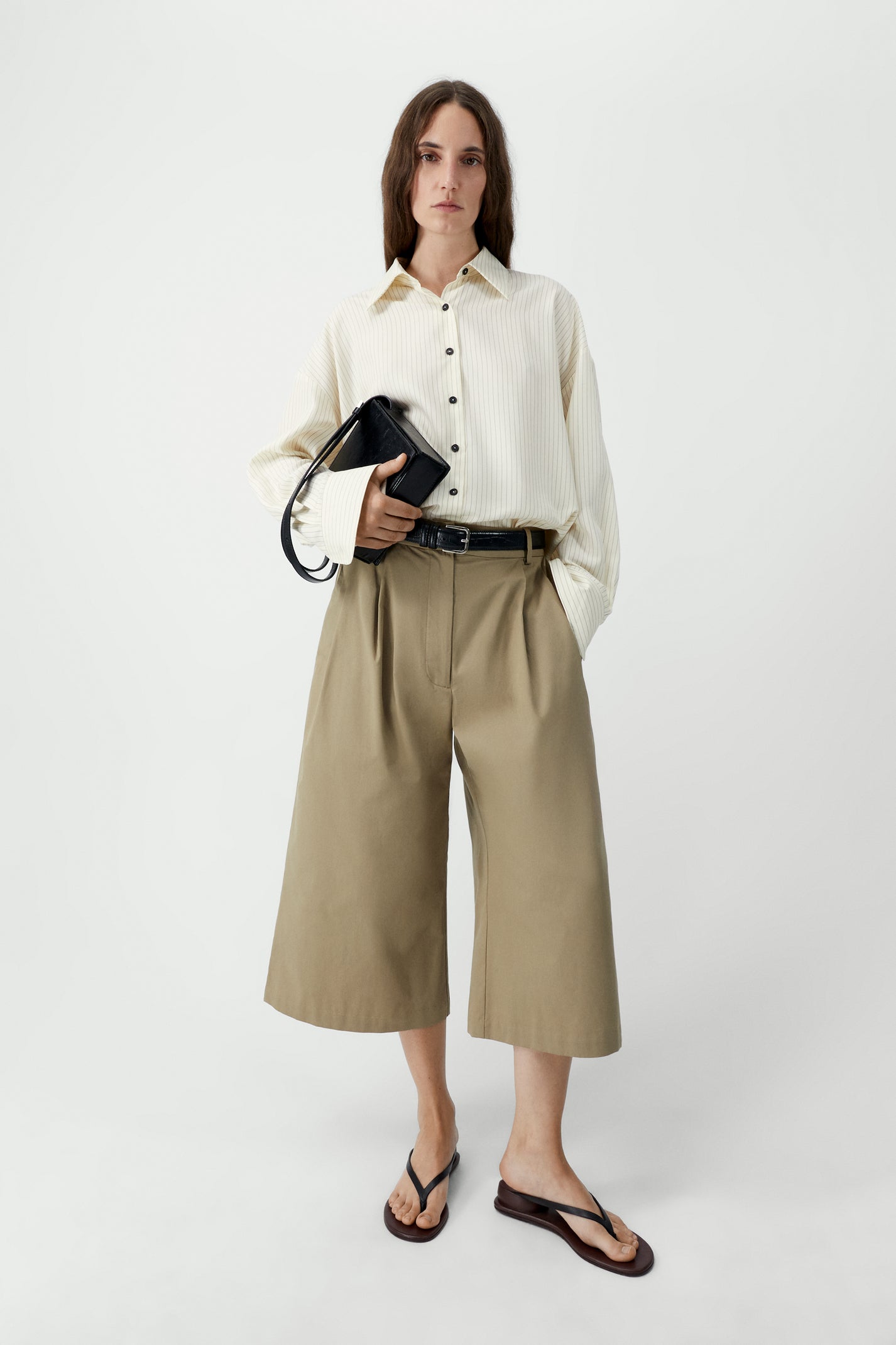 Woman wearing a white shirt and beige wide-leg pants holding a black clutch against a white background