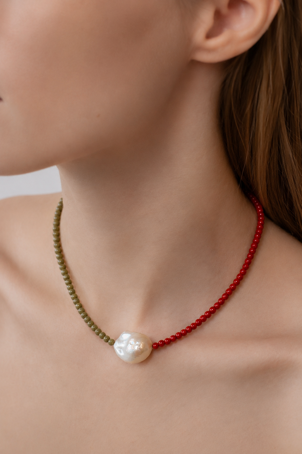 close up of a person wearing a read and sage coloured necklace with a central baroque pearl